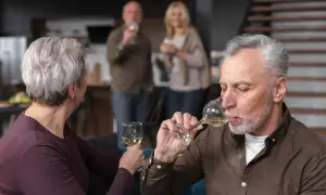 Starszy mężczyzna pije wino, zastanawiając się, czy alkoholizm jest dziedziczny. W tle para wznosi toast.