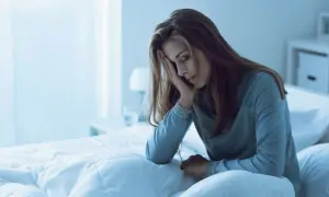Kobieta w łóżku, z ręką na czole, wygląda na zmęczoną i zmartwioną. Zastanawia się, ile zaraza się grypa.