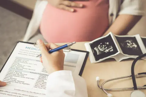 Lekarz wypełnia dokumentację medyczną pacjentki w ciąży, na stole leżą zdjęcia USG płodu i stetoskop. Badanie może wykryć mięśniaki macicy.