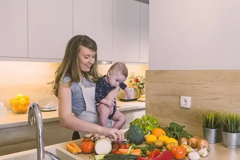 Mama z dzieckiem w kuchni, przygotowuje zdrowe jedzenie. Czosnek i inne warzywa na blacie, idealne dla karmienia piersią.