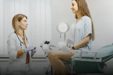 Lekarka w rękawiczkach rozmawia z pacjentką siedzącą na fotelu ginekologicznym. To jej pierwsza wizyta u ginekologa, czuje się trochę zestresowana, ale lekarz ją uspokaja.