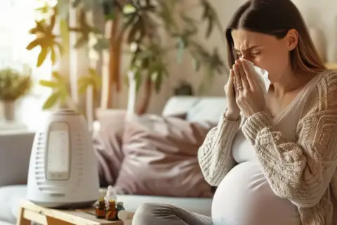 Ciężarna kobieta z chusteczką przy nosie szuka informacji: co na ból gardła w ciąży. Obok nawilżacz powietrza i buteleczki.