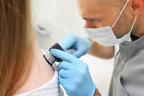 Dermatolog w rękawiczkach bada pieprzyki pacjentki dermatoskopem, kluczowy krok w tym, jak wykryć raka skóry.