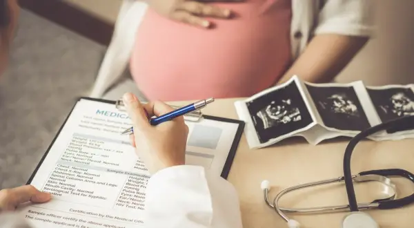 Lekarz wypełnia dokumentację medyczną pacjentki w ciąży, na stole leżą zdjęcia USG płodu i stetoskop. Badanie może wykryć mięśniaki macicy.