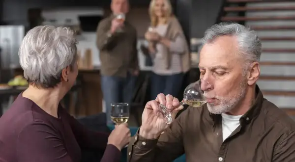 Starszy mężczyzna pije wino, zastanawiając się, czy alkoholizm jest dziedziczny. W tle para wznosi toast.
