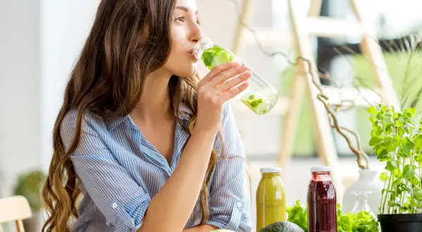 Kobieta pije wodę z ogórkiem i miętą, wspierając szybki detoks odchudzający. Na stole świeże warzywa i owoce.