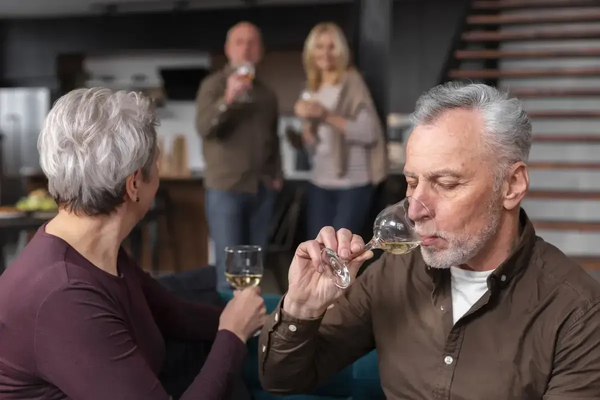 Starszy mężczyzna pije wino, zastanawiając się, czy alkoholizm jest dziedziczny. W tle para wznosi toast.