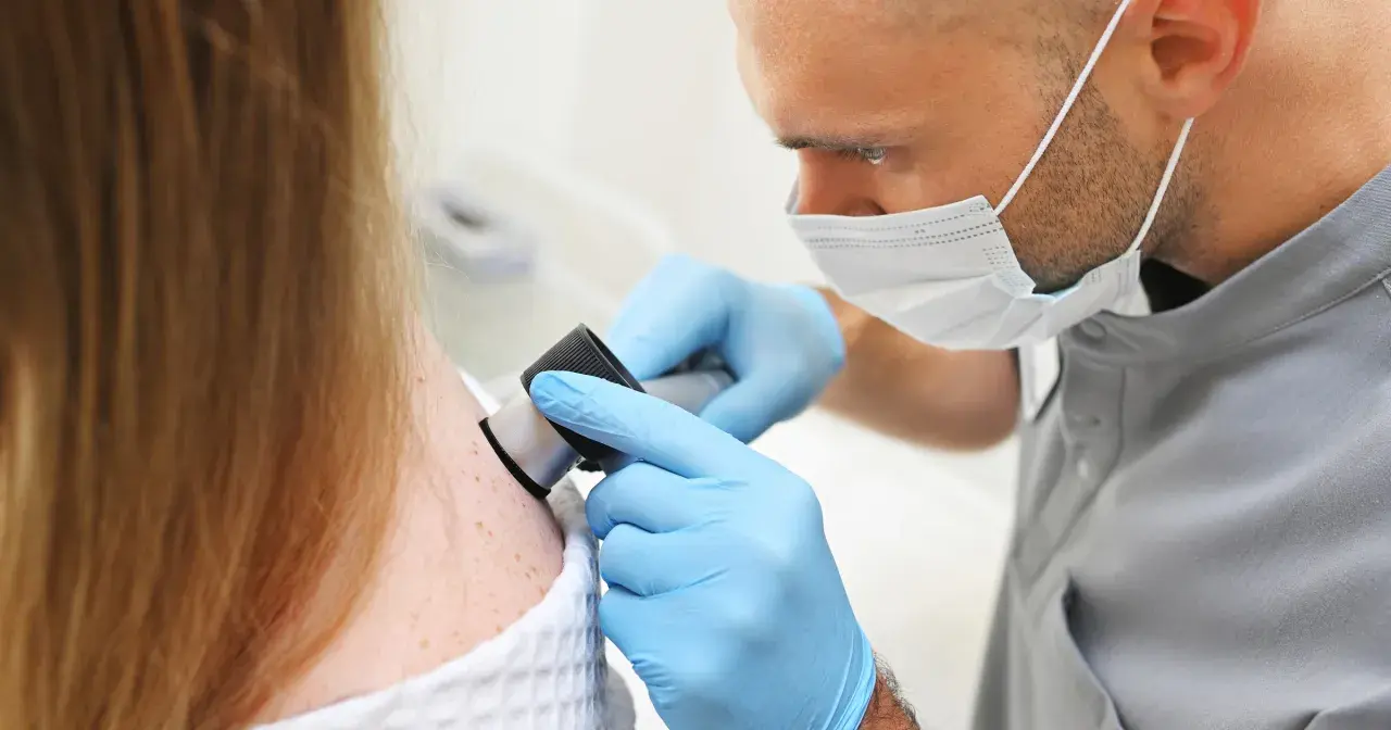 Dermatolog w rękawiczkach bada pieprzyki pacjentki dermatoskopem, kluczowy krok w tym, jak wykryć raka skóry.