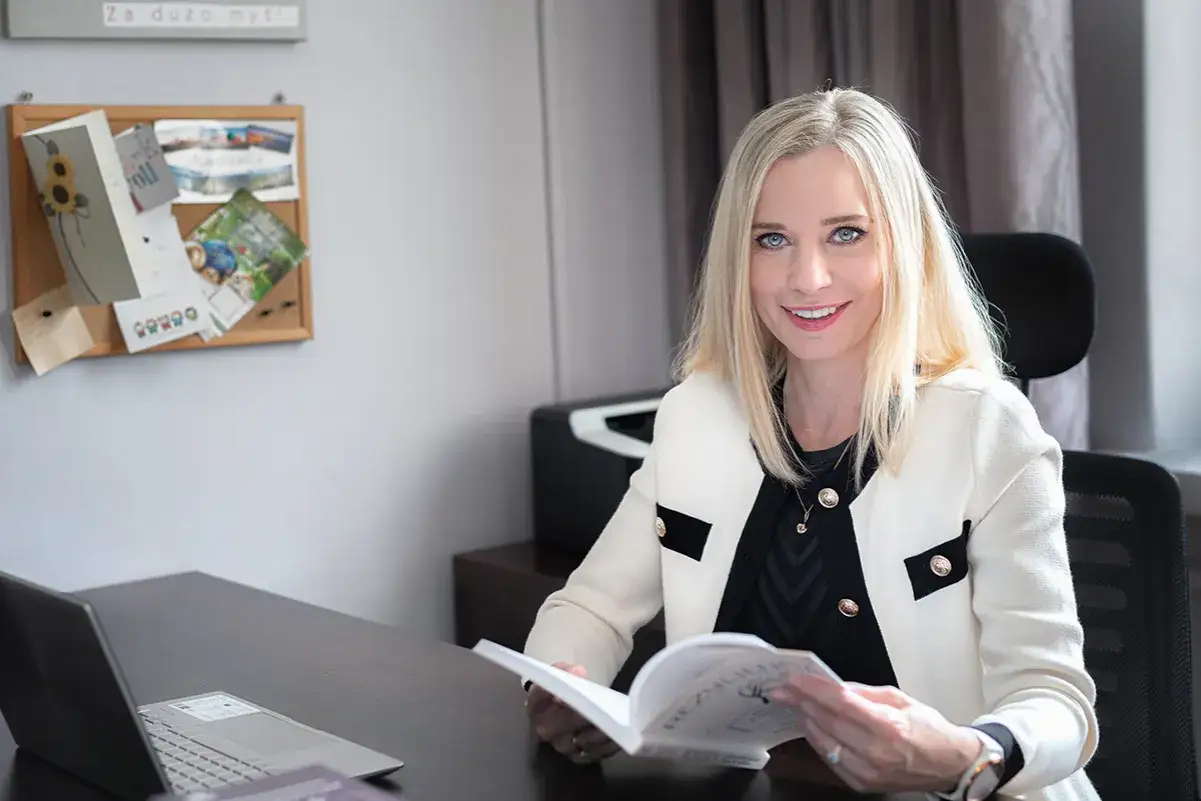 Uśmiechnięta terapeutka poznawczo-behawioralna z blond włosami siedzi przy biurku, czytając książkę.